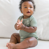 An infant wearing a sage green organic onesie, sitting on a white linen bed.